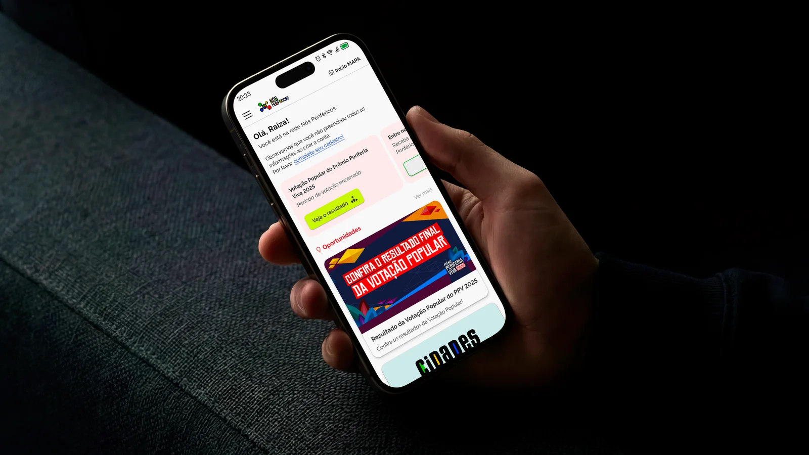 Celular na mão mostrando a tela inicial do Nós Periféricos, com saudação, aviso para completar o cadastro, destaque da Votação Popular e oportunidades